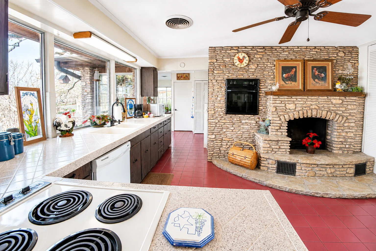 Open-plan kitchen and living area with brick fireplace, red tile floors, dark cabinets, and rooster decor at Scurlock Farms