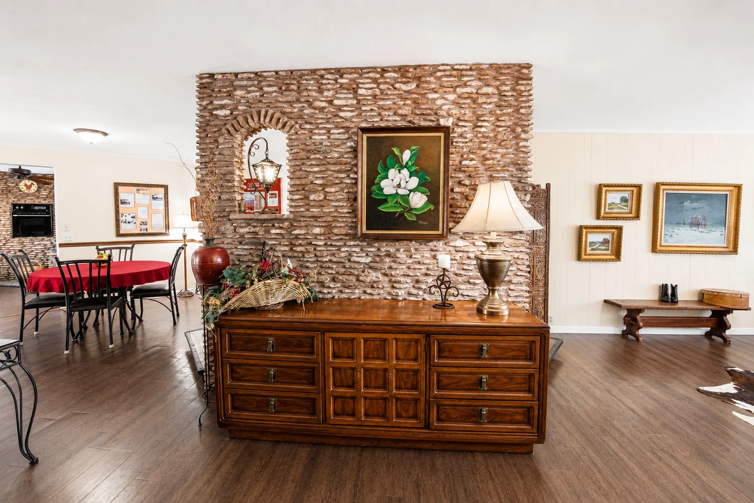 Living room with exposed brick accent wall, framed botanical art, wooden dresser, and red-tableclothed dining area at